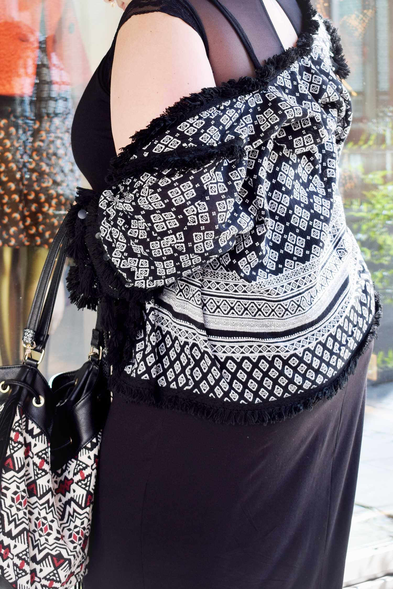 Nahaufnahme eines Oberkörpers mit schwarz-weiß gemustertem Fransen-Top über einem schwarzen Kleid, dazu eine gemusterte Tasche