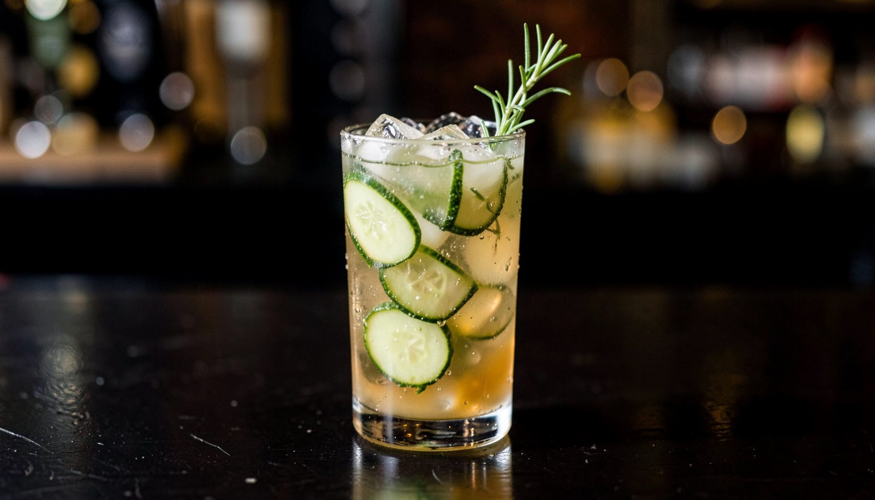 A tall glass cocktail with amber liquid, ice, cucumber slices, and a rosemary sprig garnish, set on a dark bar counter
