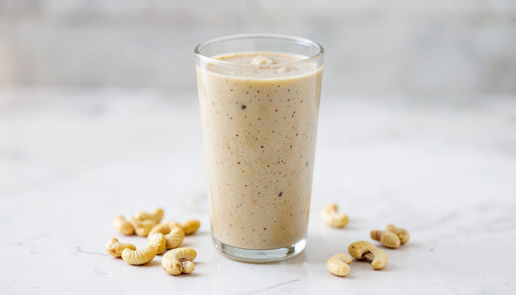 A glass of beige-colored smoothie surrounded by scattered raw cashew nuts on a white surface